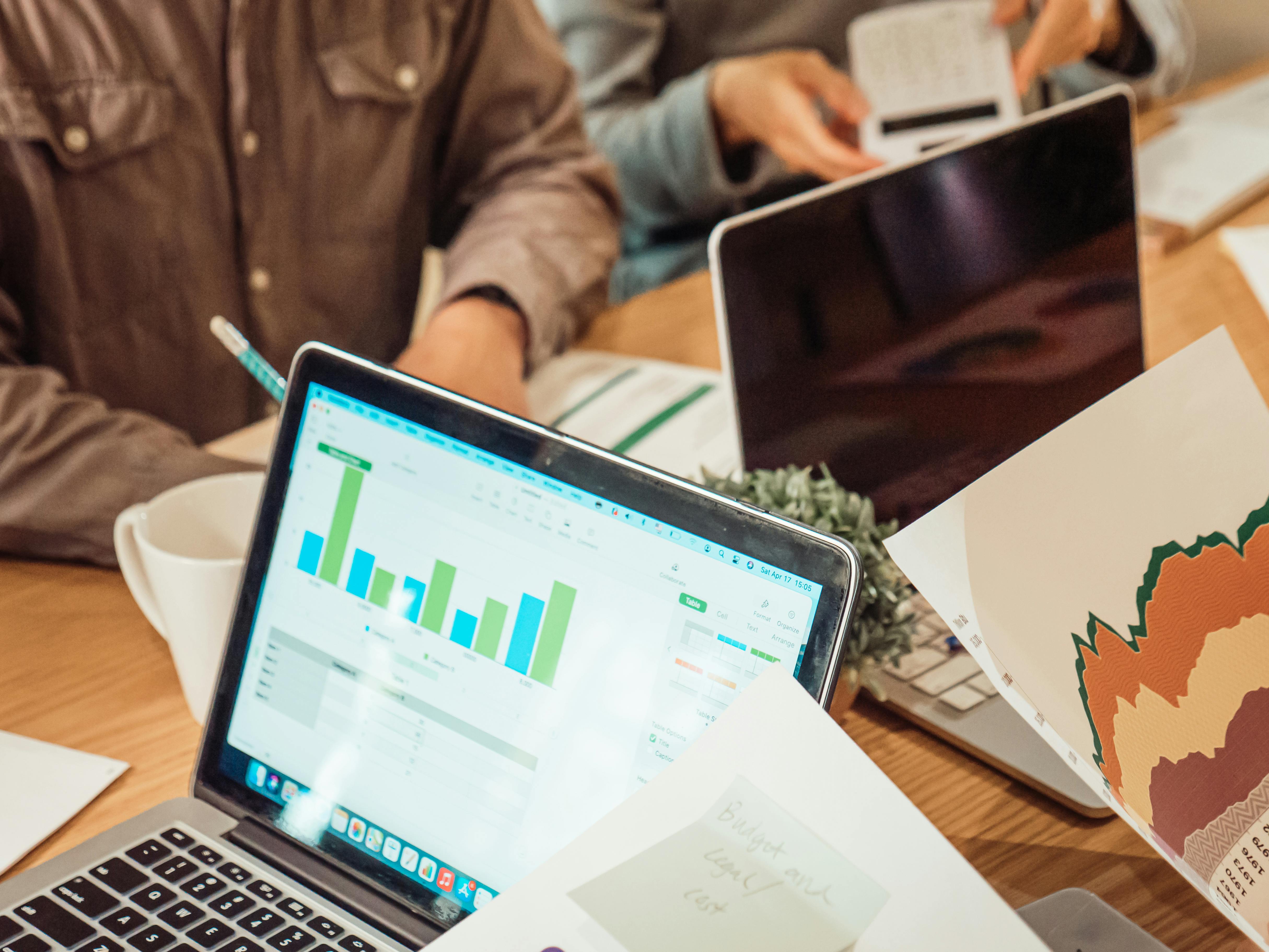 Close-up of a laptop screen displaying business charts and graphs during a meeting, with documents and other laptops in the background.